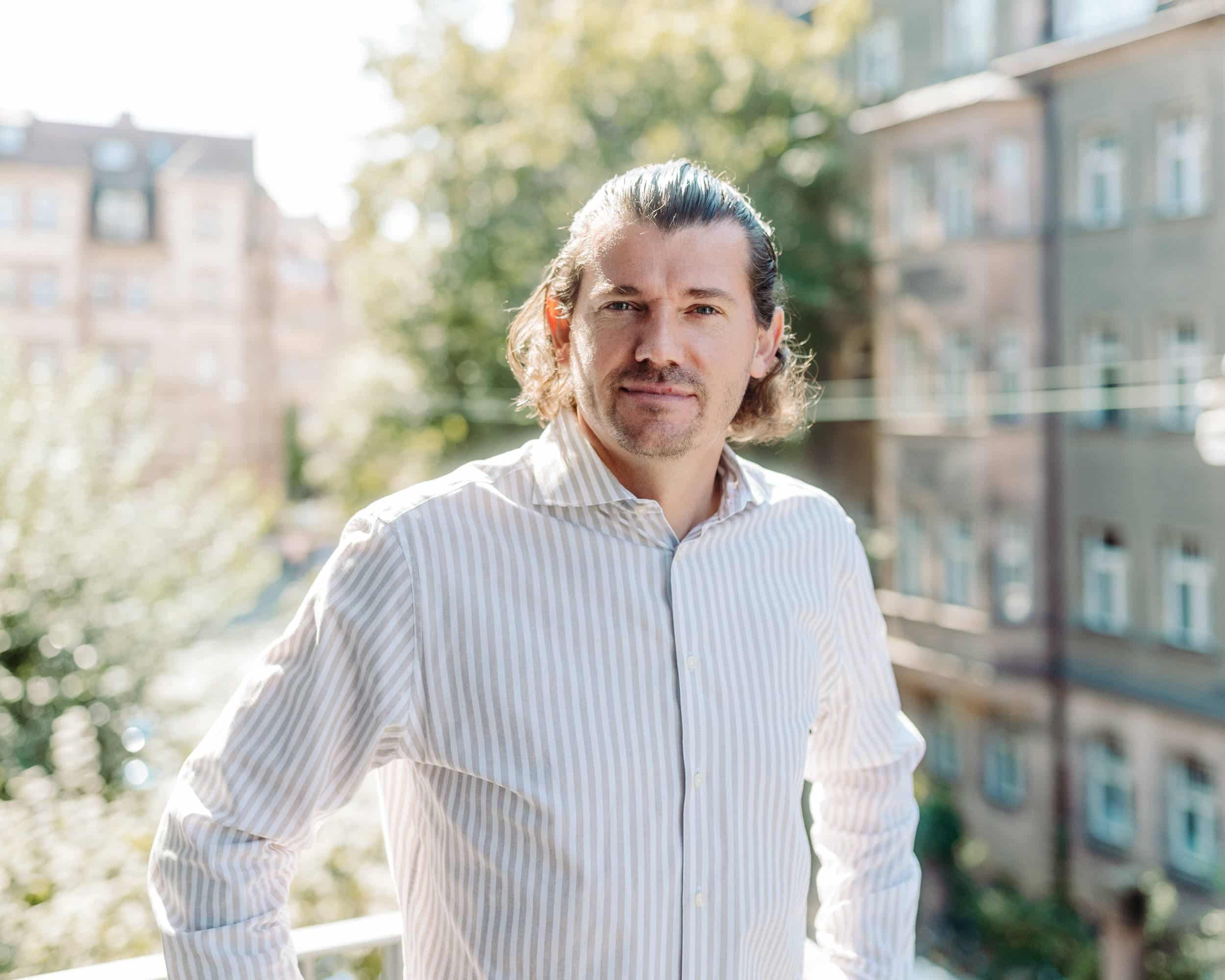 Toni steht lächelnd auf einem Balkon, der von Sonnenlicht durchflutet wird. Die entspannte Pose und die Sonne reflektieren eine positive und offene Ausstrahlung.